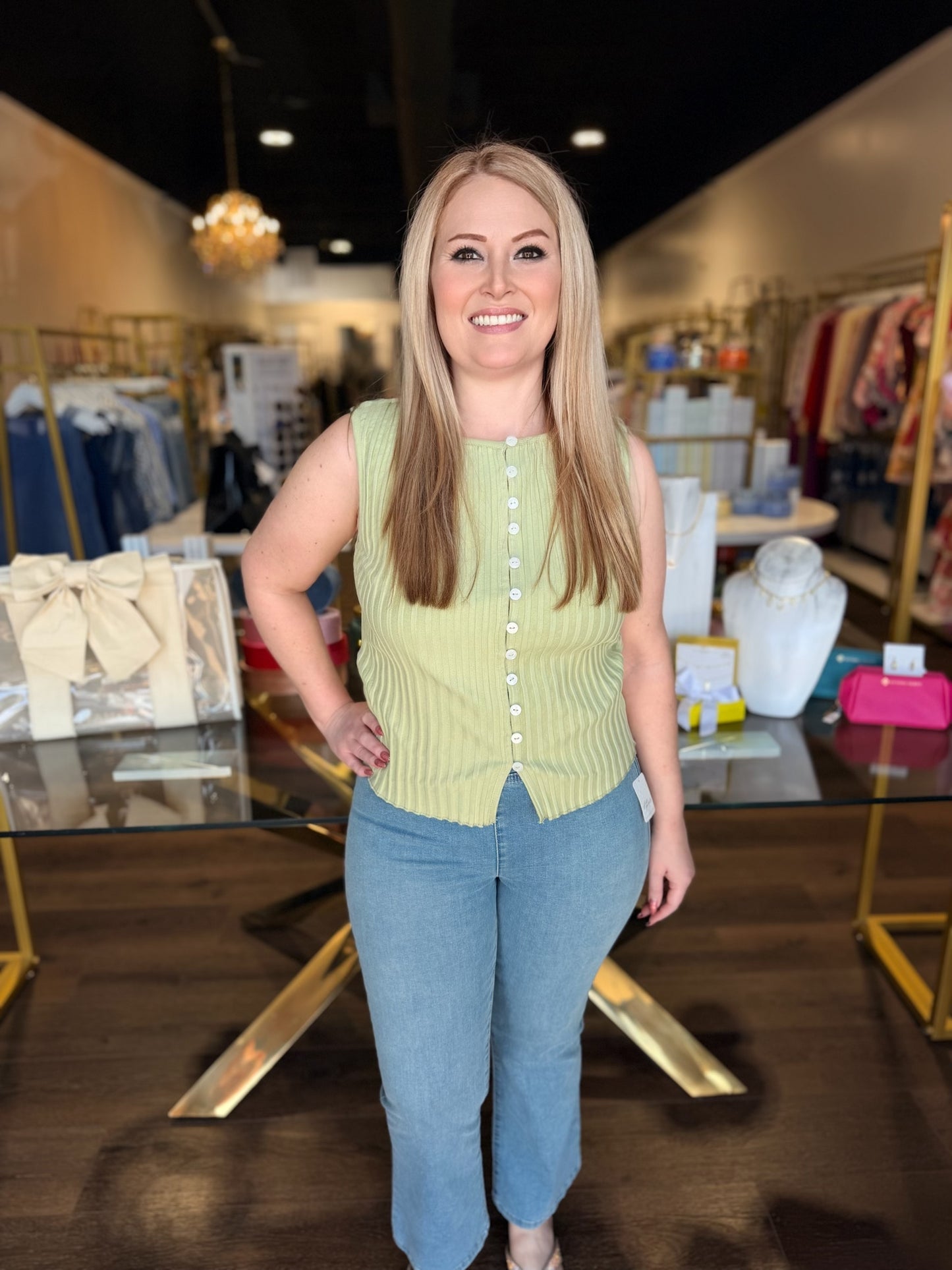 Woman standing in a clothing store wearing a green sleeveless top and blue jeans.