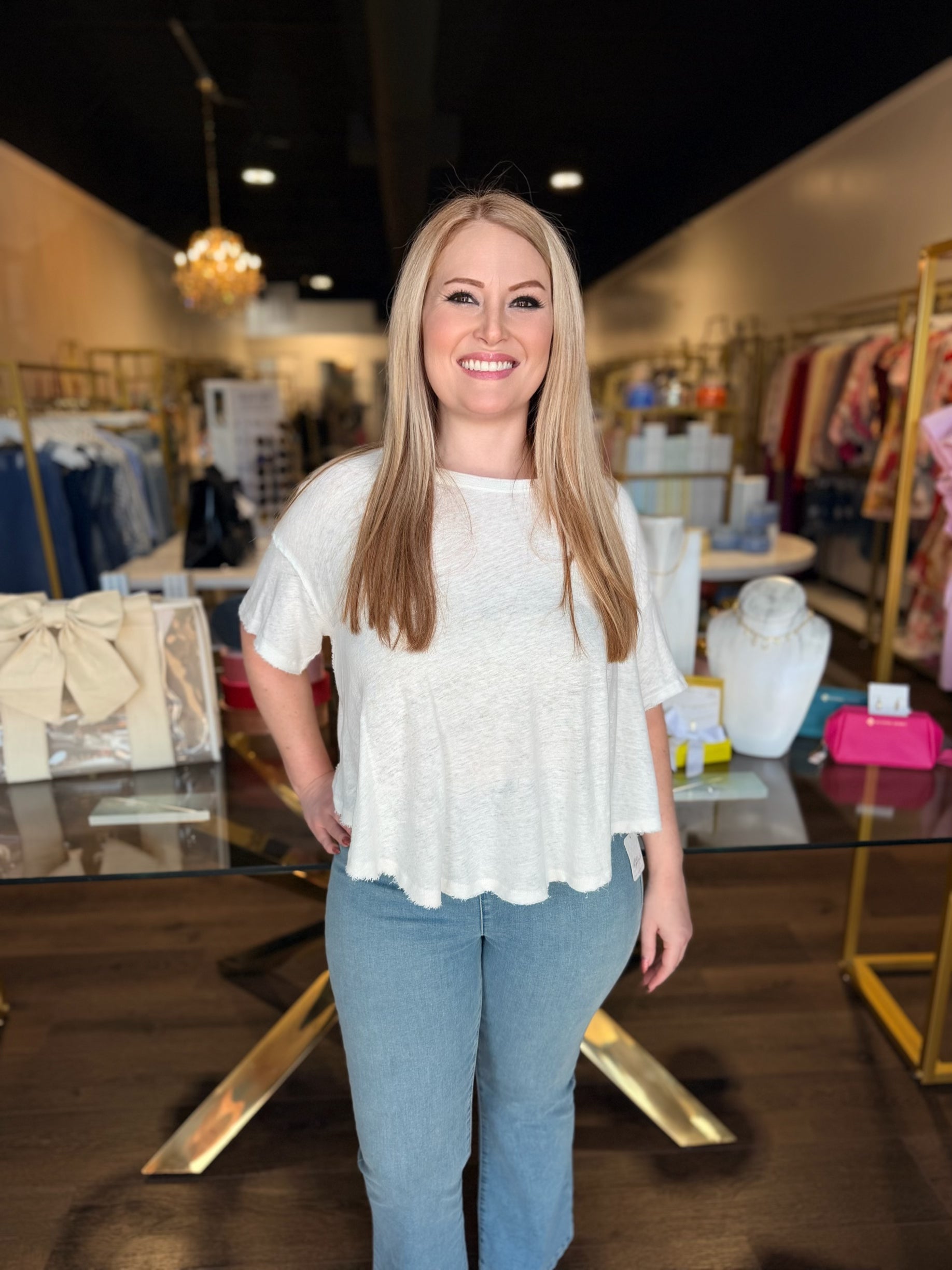 Woman in a white blouse and blue jeans standing in a store with clothing racks and displays in the background.