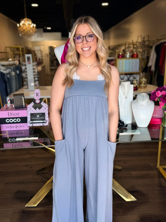 Woman wearing a light blue jumpsuit in a store setting with products and clothing racks in the background.