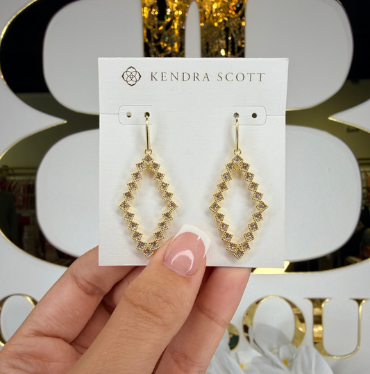 Gold earrings on a Kendra Scott card held by a hand with a decorative background.
