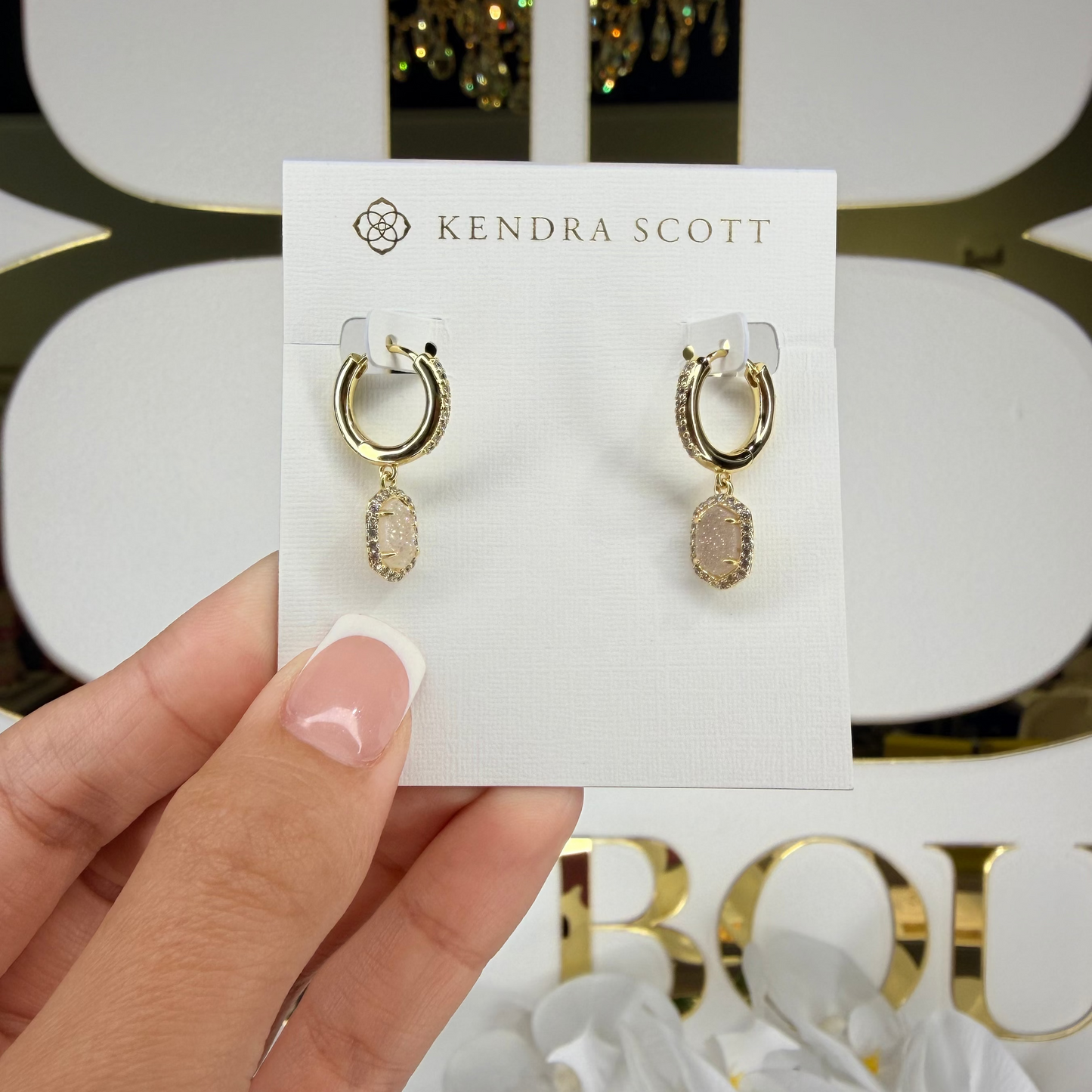 Gold hoop earrings on a Kendra Scott card held by a hand with a decorative background.