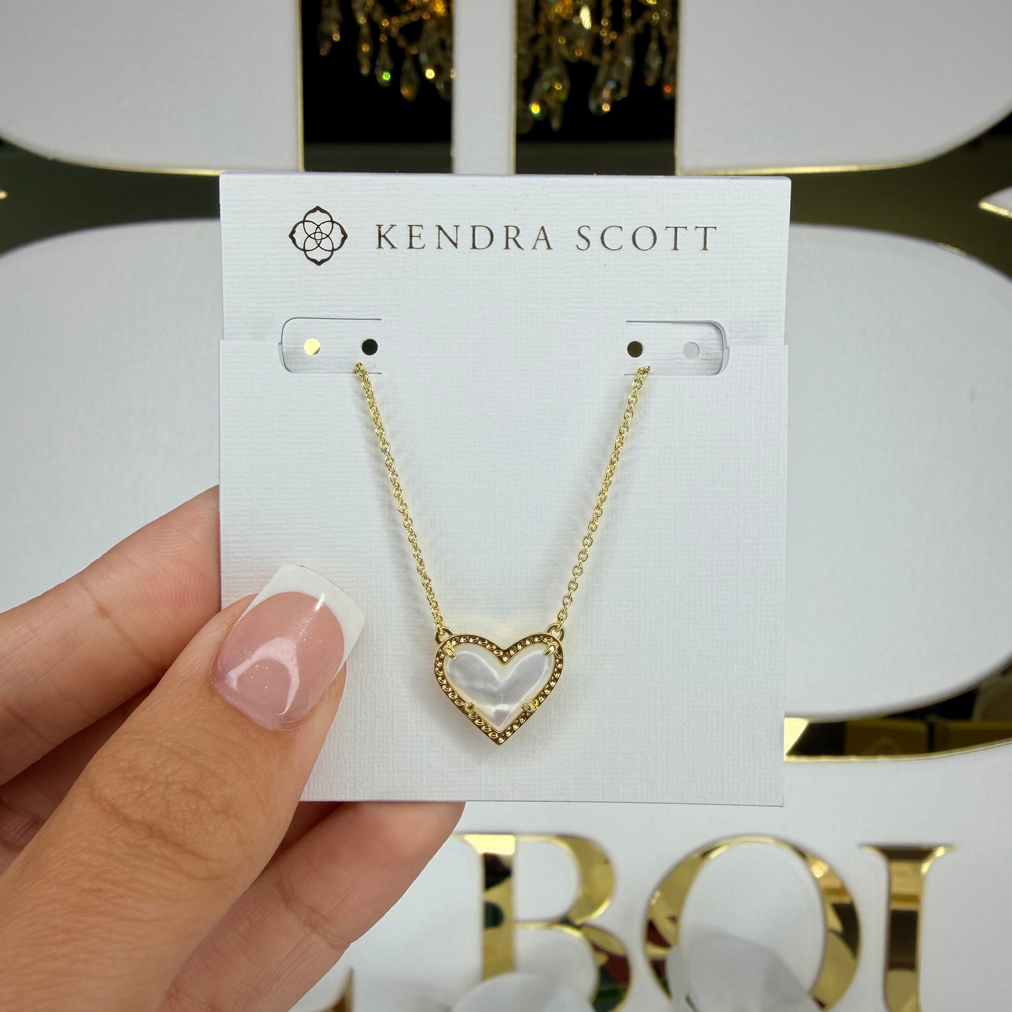 Gold heart-shaped necklace on a Kendra Scott card held by a hand with a floral background.