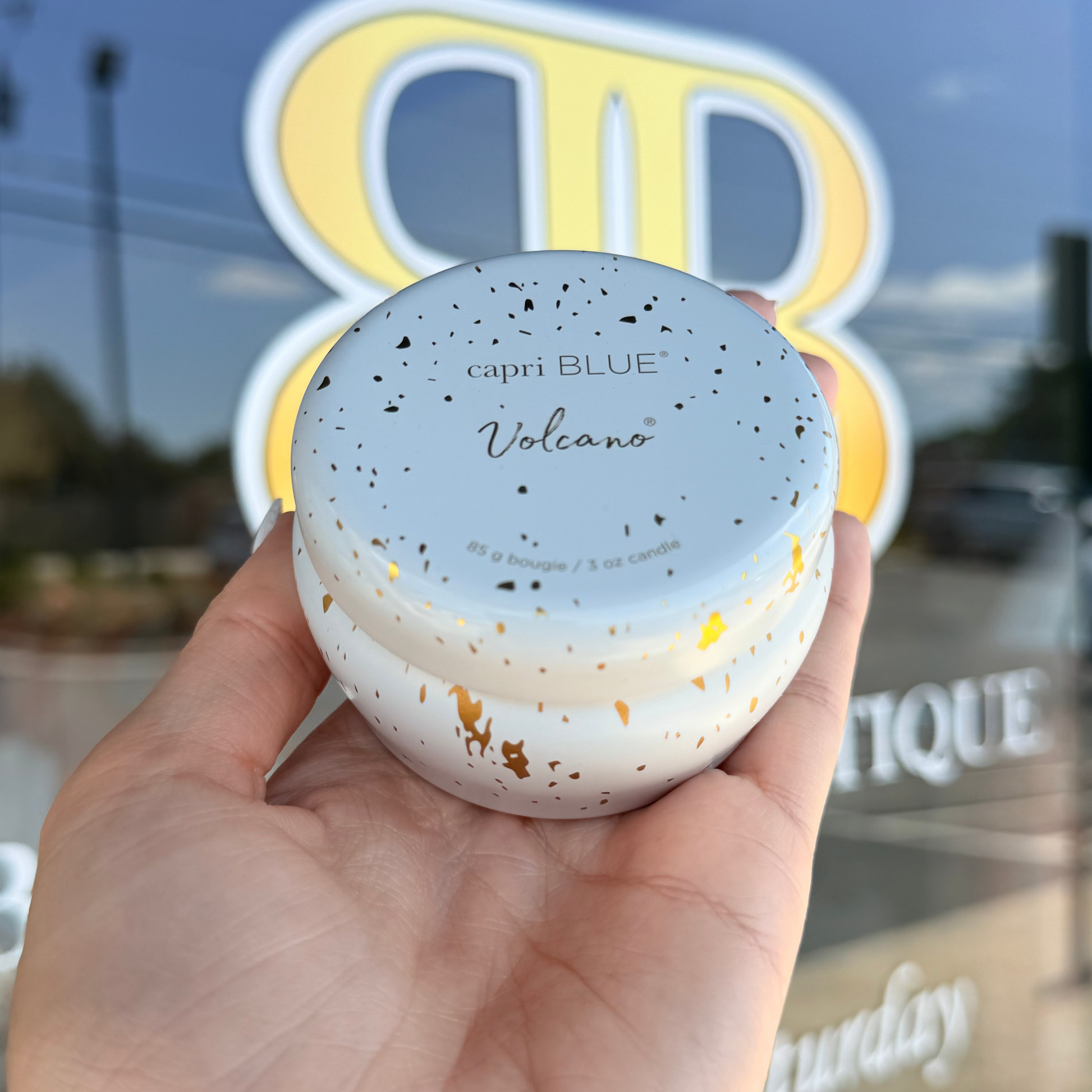 Hand holding a small container labeled 'capri BLUE Volcano' in front of a store window with 'BR' branding.