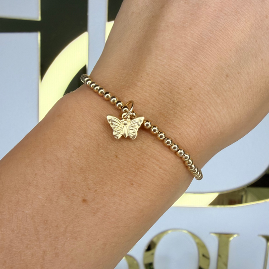 Gold bracelet with a butterfly charm on a wrist, blurred background with decorative elements.
