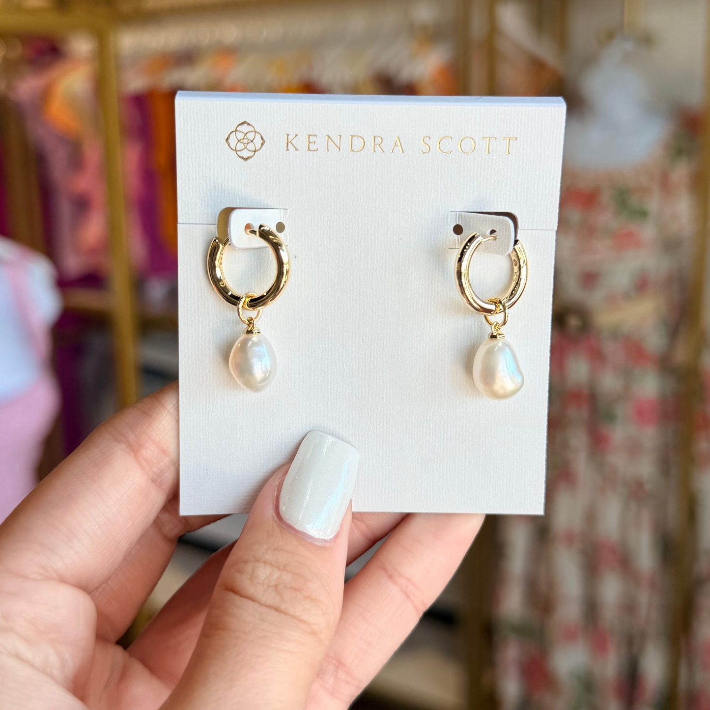 Kendra Scott earrings on a card held by a hand with a blurred store background.