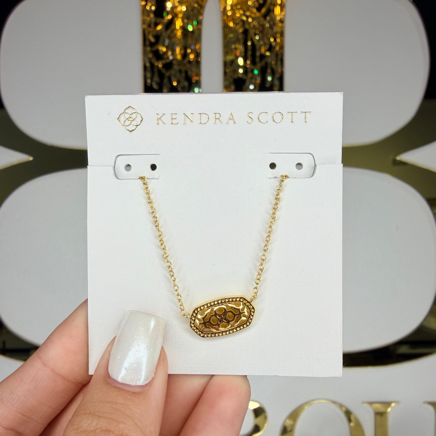 Gold necklace on a Kendra Scott card held by a hand with a decorative background.
