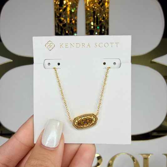 Gold necklace on a Kendra Scott card held by a hand with a decorative background.