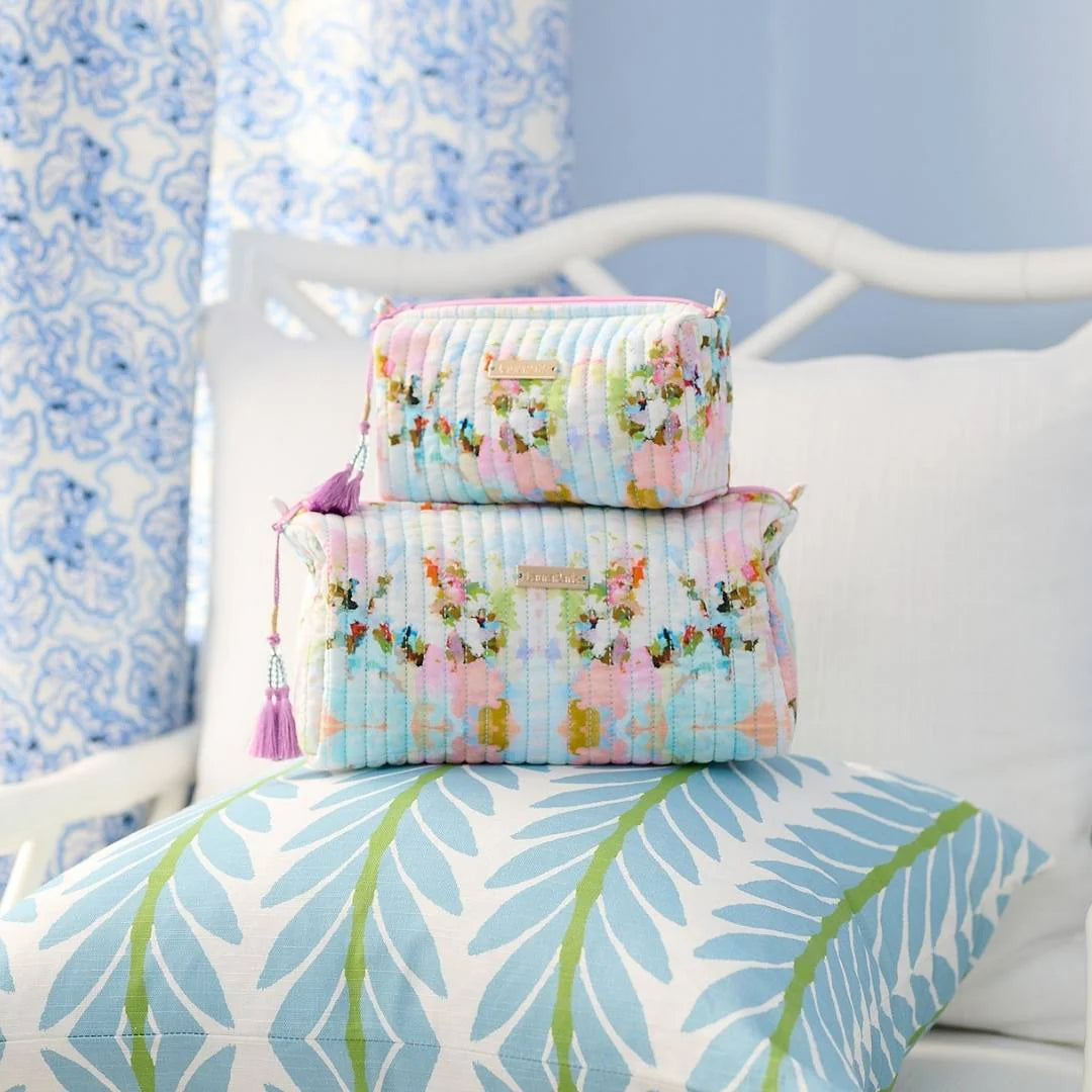 Stack of floral-patterned Laura Park cosmetic bags on a bed with a blue and white curtain in the background