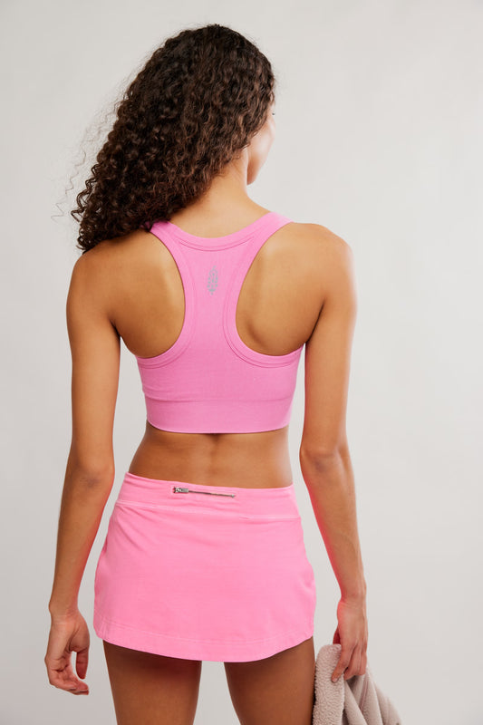 A woman shown from the back wearing a bright pink athletic crop top and matching pink tennis skirt against a light gray studio background.