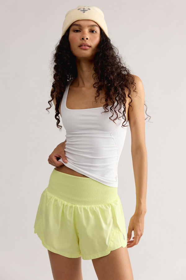 A woman modeling a white ribbed tank top and light yellow-green athletic shorts against a plain light gray studio background. She is also wearing a cream beanie.