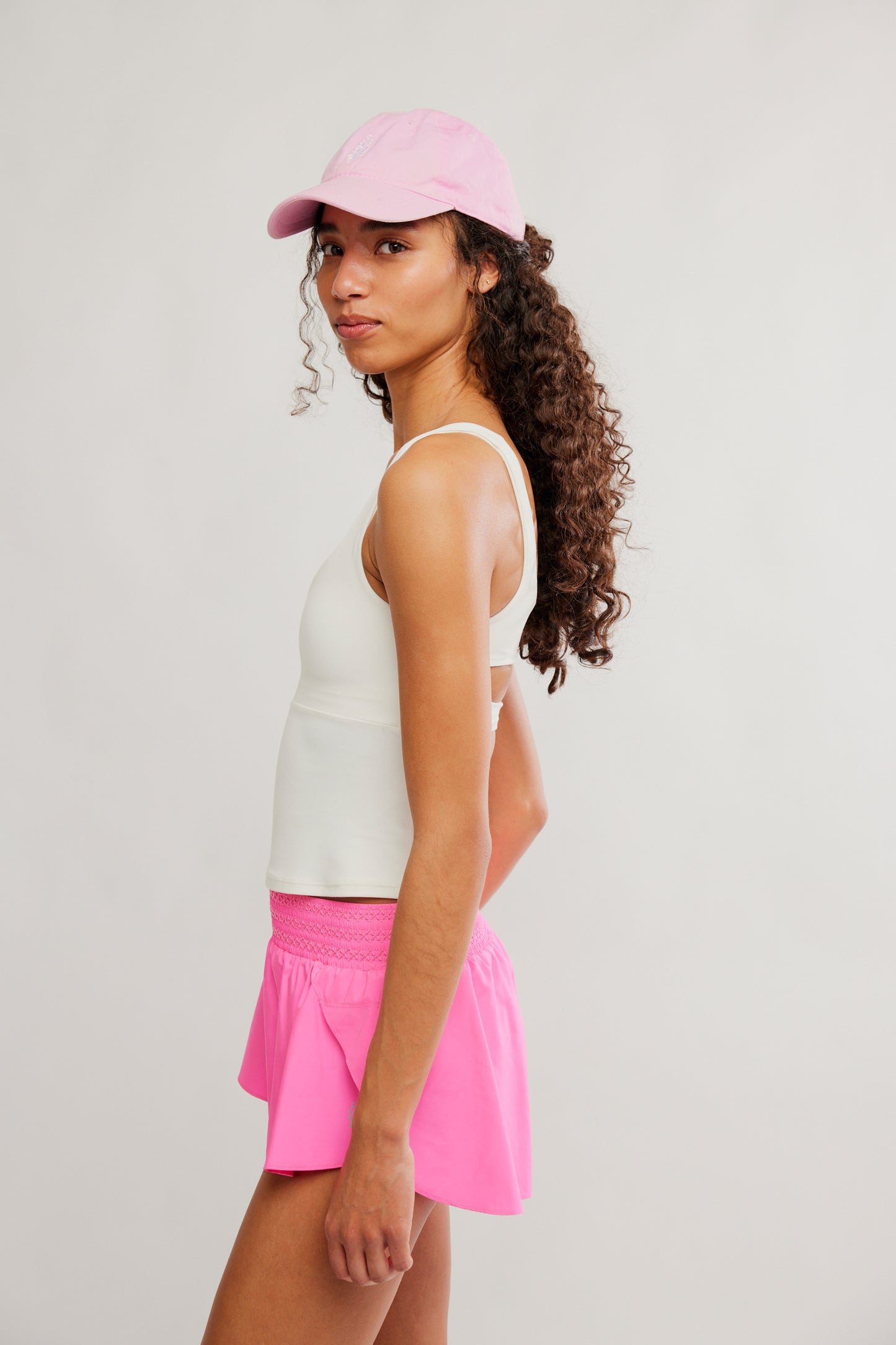 Woman wearing a white tank top, pink shorts, and pink cap on a light gray background