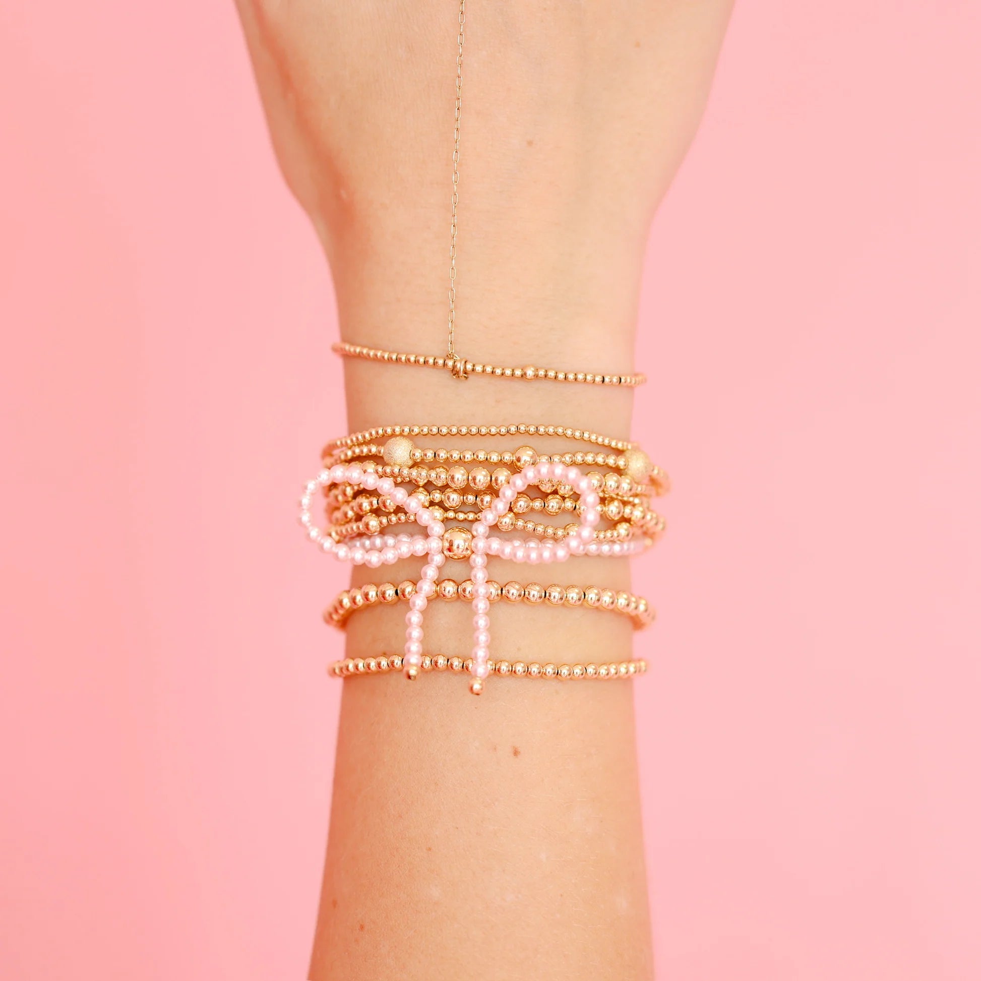 A stack of delicate beaded bracelets on a person's wrist, featuring gold-toned bead strands, white pearl-like beads, and a bow-shaped charm, set against a solid pink background.