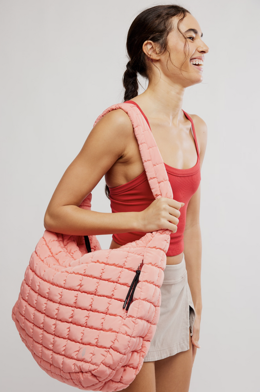 A woman wearing a red tank top and white shorts carries a large, quilted pink shoulder bag with a wide strap against a light gray studio background.