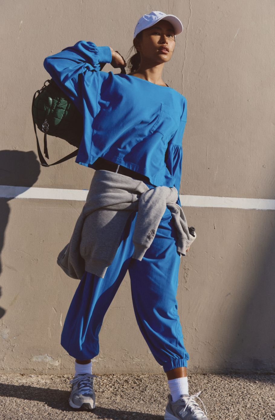 A woman wearing a bright blue cropped long-sleeve top and matching blue jogger pants, with a gray sweatshirt tied around her waist, a white cap, and sneakers, standing against a beige wall.