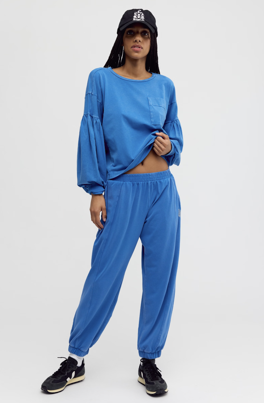 A model wearing a matching blue two-piece lounge set consisting of a cropped long-sleeve top with balloon sleeves and matching jogger pants, styled with a black baseball cap, sunglasses, and black sneakers against a light gray studio background.