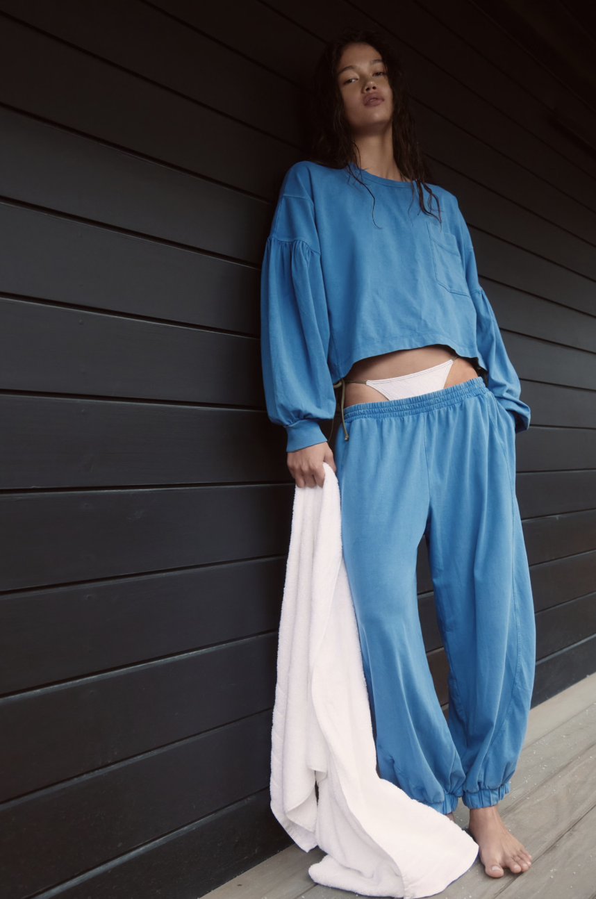 A woman stands barefoot against a dark wood-paneled wall wearing a matching blue lounge set consisting of a cropped long-sleeve top and wide-leg pants, while holding a white garment drape.