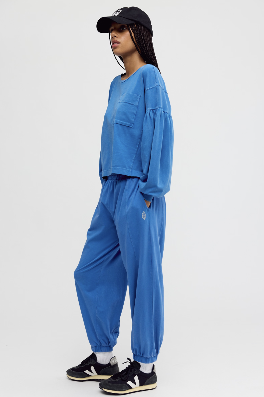 A model wearing a matching blue lounge set consisting of a cropped long-sleeve top and jogger pants, styled with a black baseball cap, white socks, and black sneakers against a light gray background.