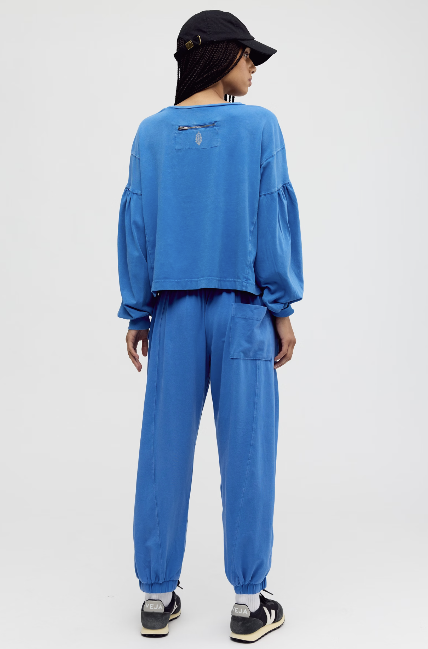 A model shown from the back wearing a matching blue two-piece lounge set consisting of a cropped long-sleeve top with balloon sleeves and matching jogger pants, paired with a black cap and sneakers.