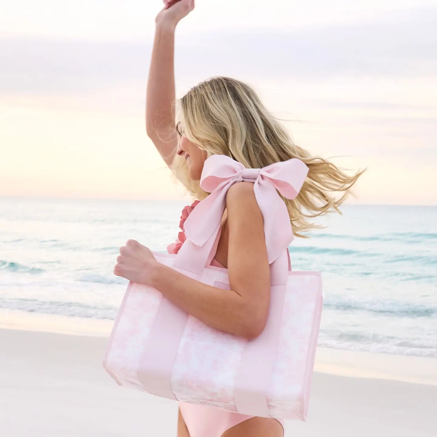 Woman in a pink dress with a large bow standing on a beach at sunset.
