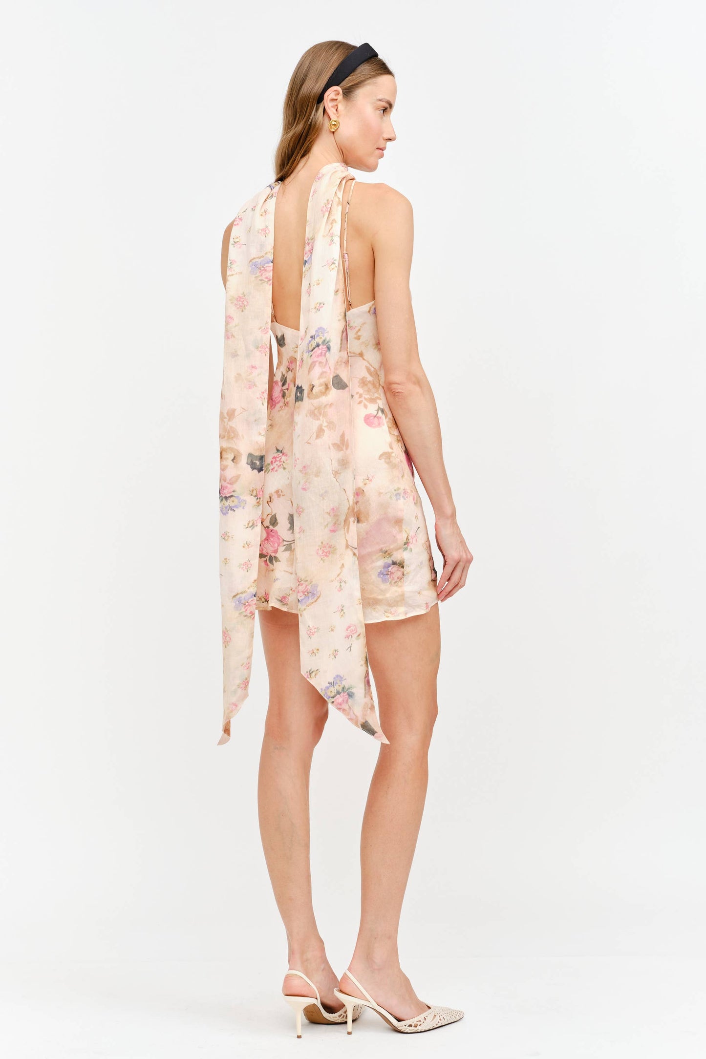 A woman modeling a sleeveless, high-low floral dress with a draped back, shown from the back against a plain white studio background.