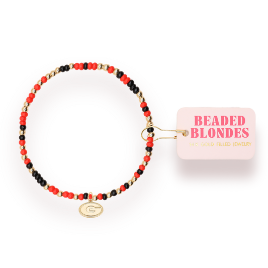A beaded stretch bracelet with red, black, and gold beads, featuring a gold-toned 'G' initial charm. It is shown with its pink paper tag that reads 'BEADED BLONDES'.