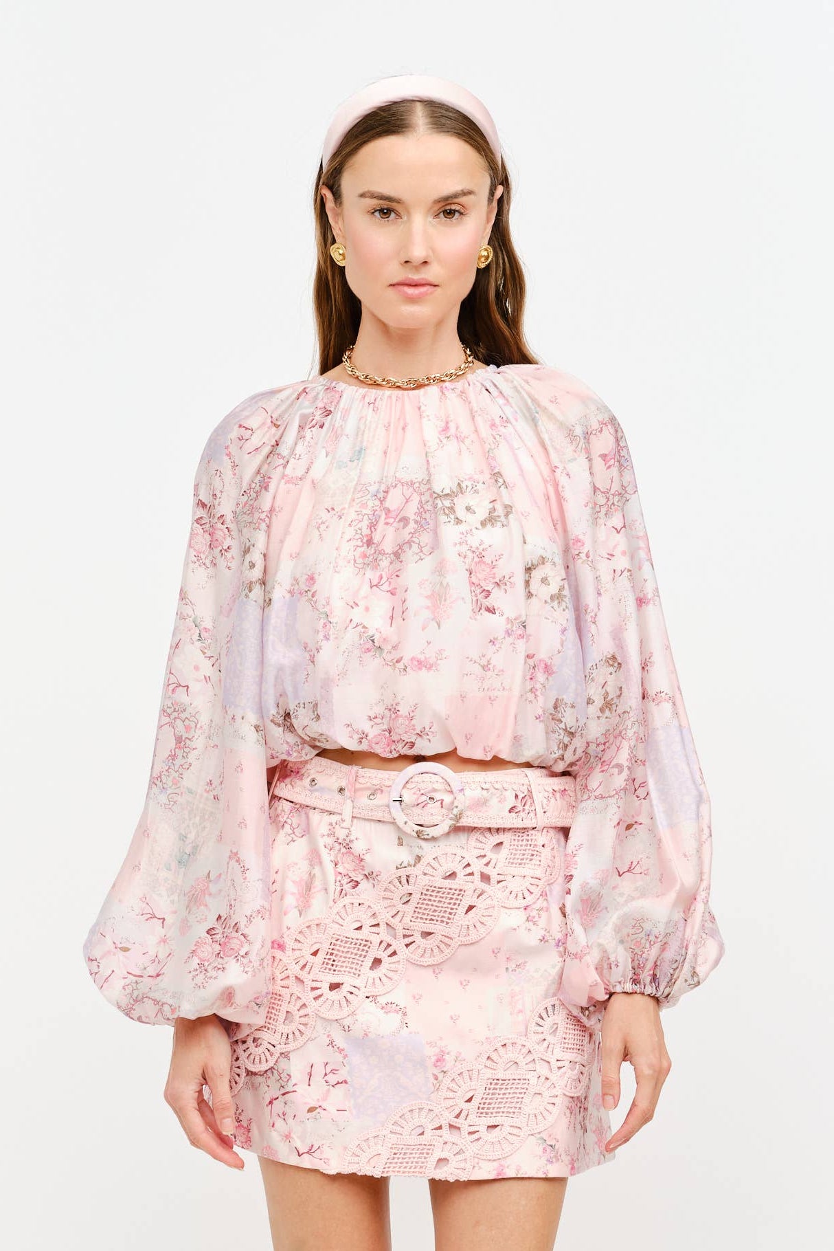 A model wearing a light pink floral two-piece set consisting of a long-sleeve cropped blouse and a matching mini skirt, paired with beige pointed-toe heels against a plain white studio background.