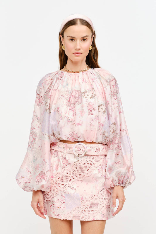 A model wearing a light pink floral two-piece set consisting of a long-sleeve cropped blouse and a matching mini skirt, paired with beige pointed-toe heels against a plain white studio background.