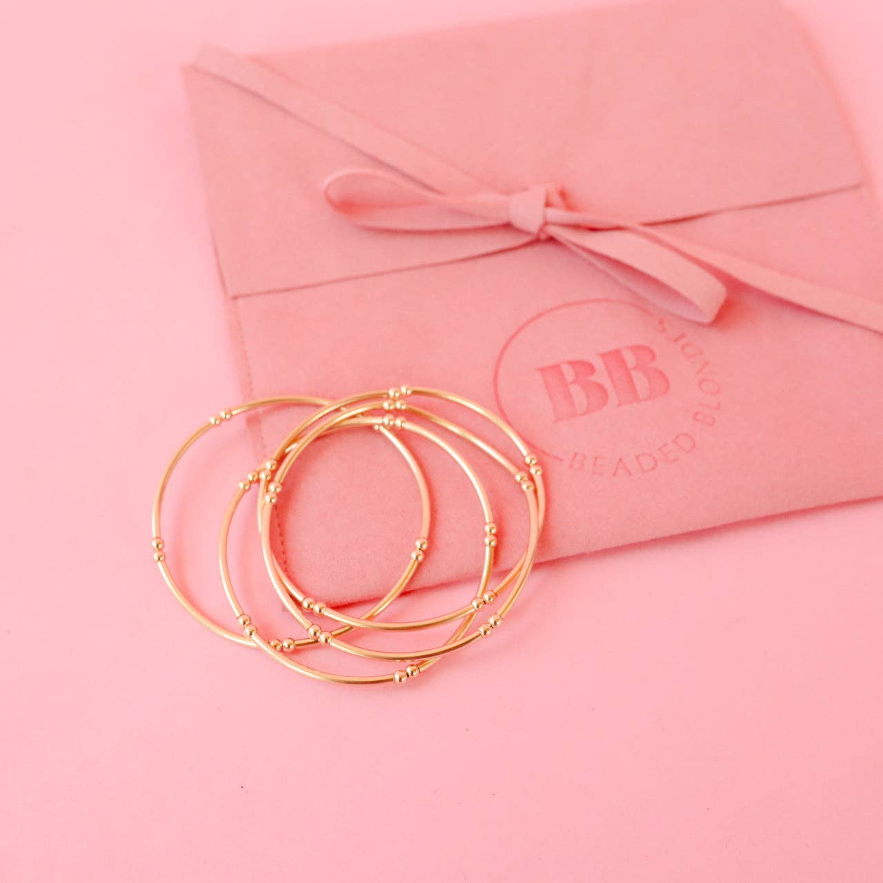 Gold bracelets on a pink background with a pink envelope featuring 'BB' branding.