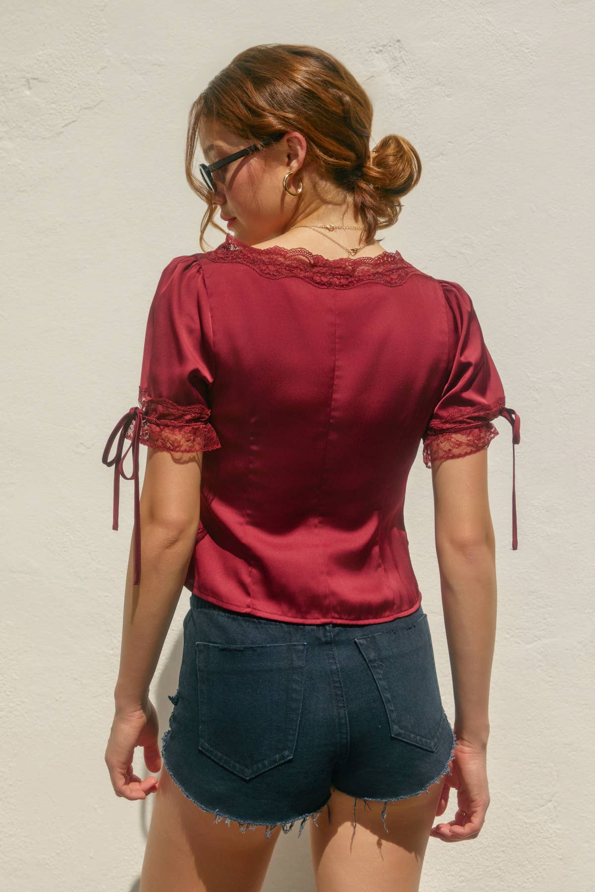 Woman wearing a red blouse with lace details and denim shorts against a light background