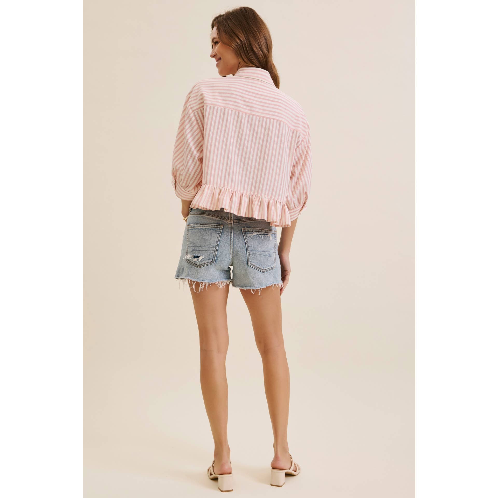 A woman is shown from behind wearing a light pink and white vertically striped ruffled blouse and light-wash denim shorts against a solid beige studio background.