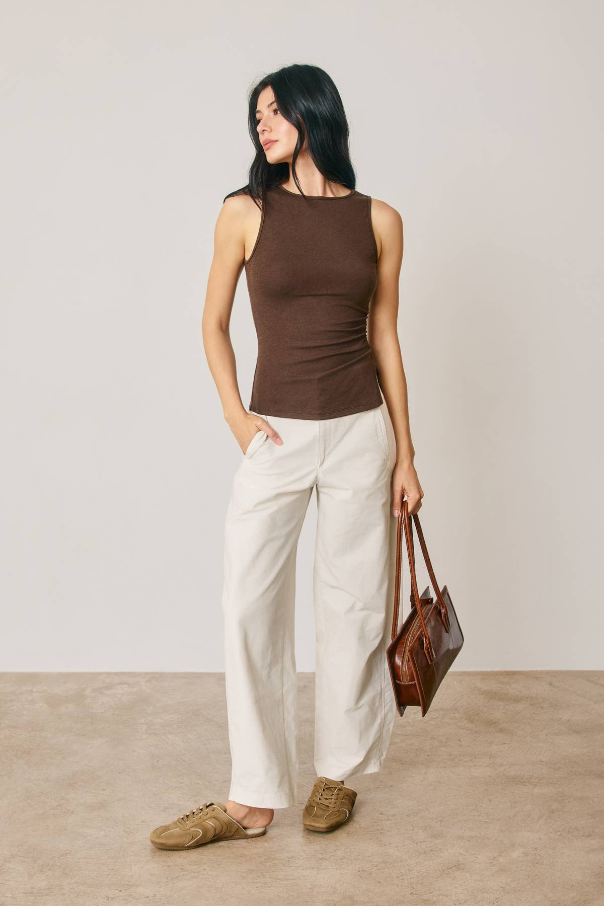 Woman wearing a brown sleeveless top and white pants, holding a brown leather bag against a plain background.