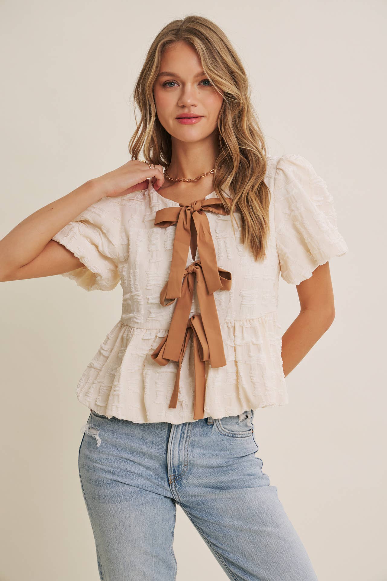 Woman wearing a white blouse with brown ribbons and light blue jeans on a plain background