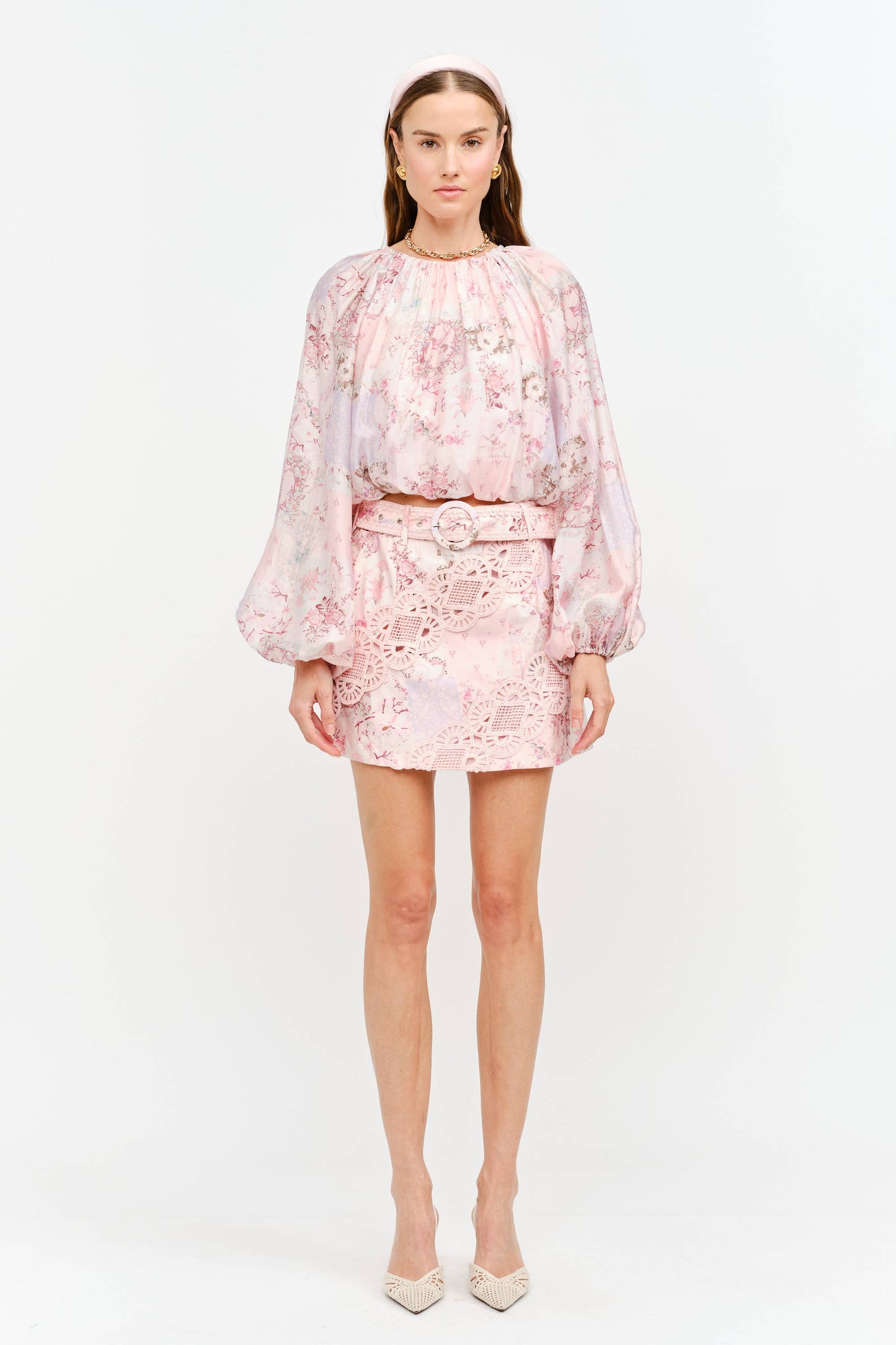 A model stands against a plain white studio background wearing a light pink floral two-piece set consisting of a long-sleeve cropped top and a matching mini skirt with a tie waist, paired with beige pointed-toe heels.