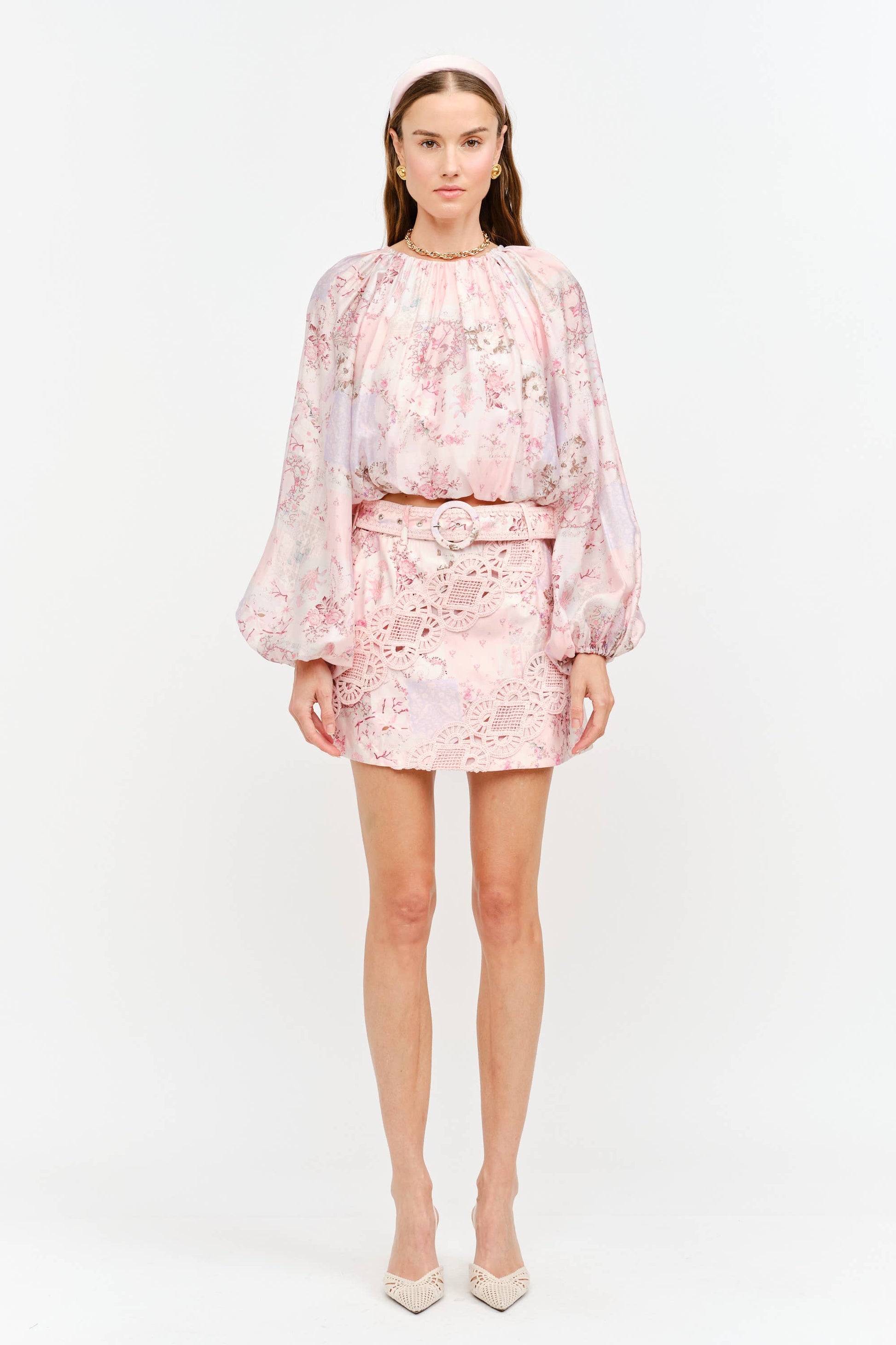 A model stands against a plain white studio background wearing a light pink floral two-piece set consisting of a long-sleeve cropped top and a matching mini skirt with a tie waist, paired with beige pointed-toe heels.