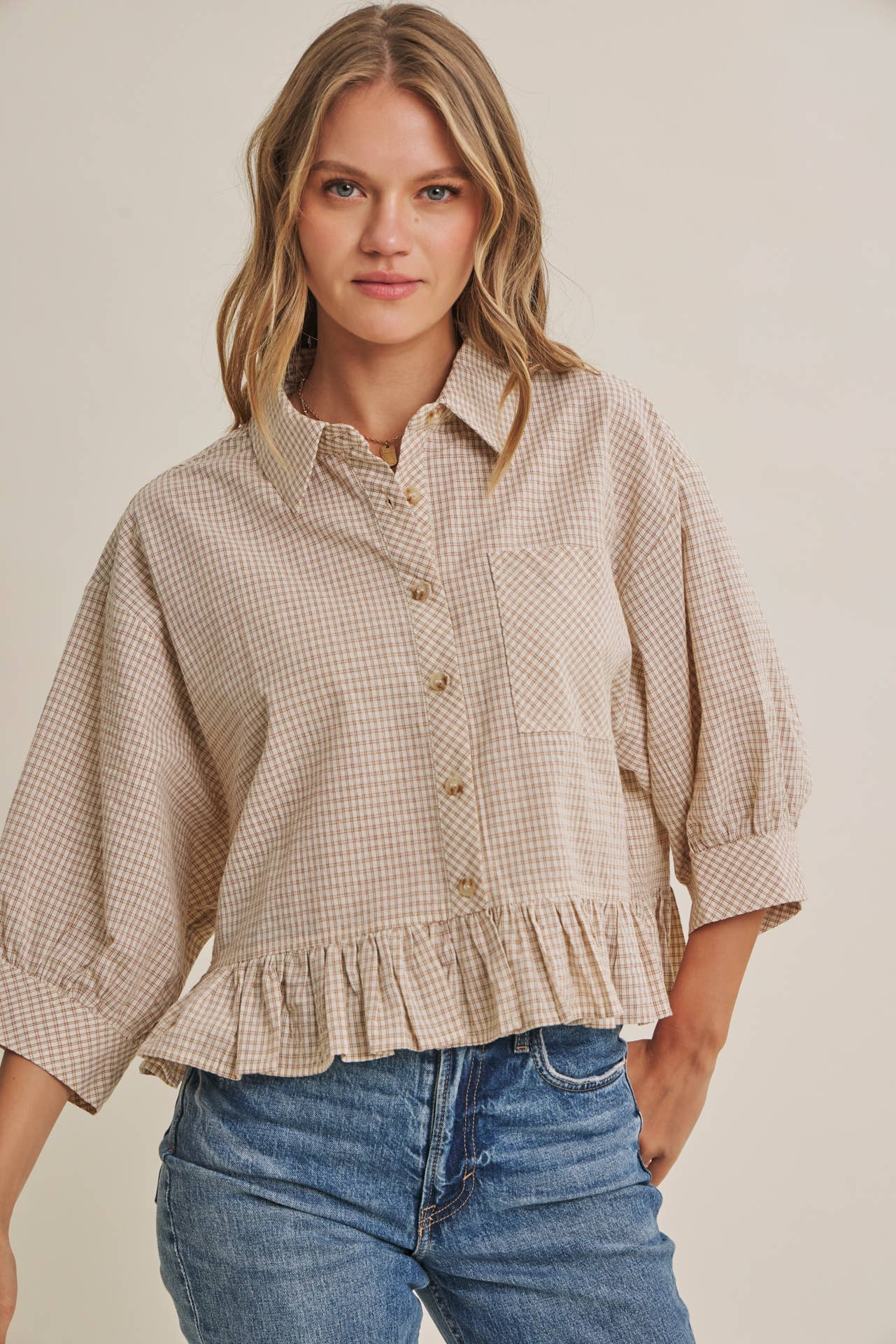 Woman wearing a beige checkered blouse with ruffled details and blue jeans on a plain background