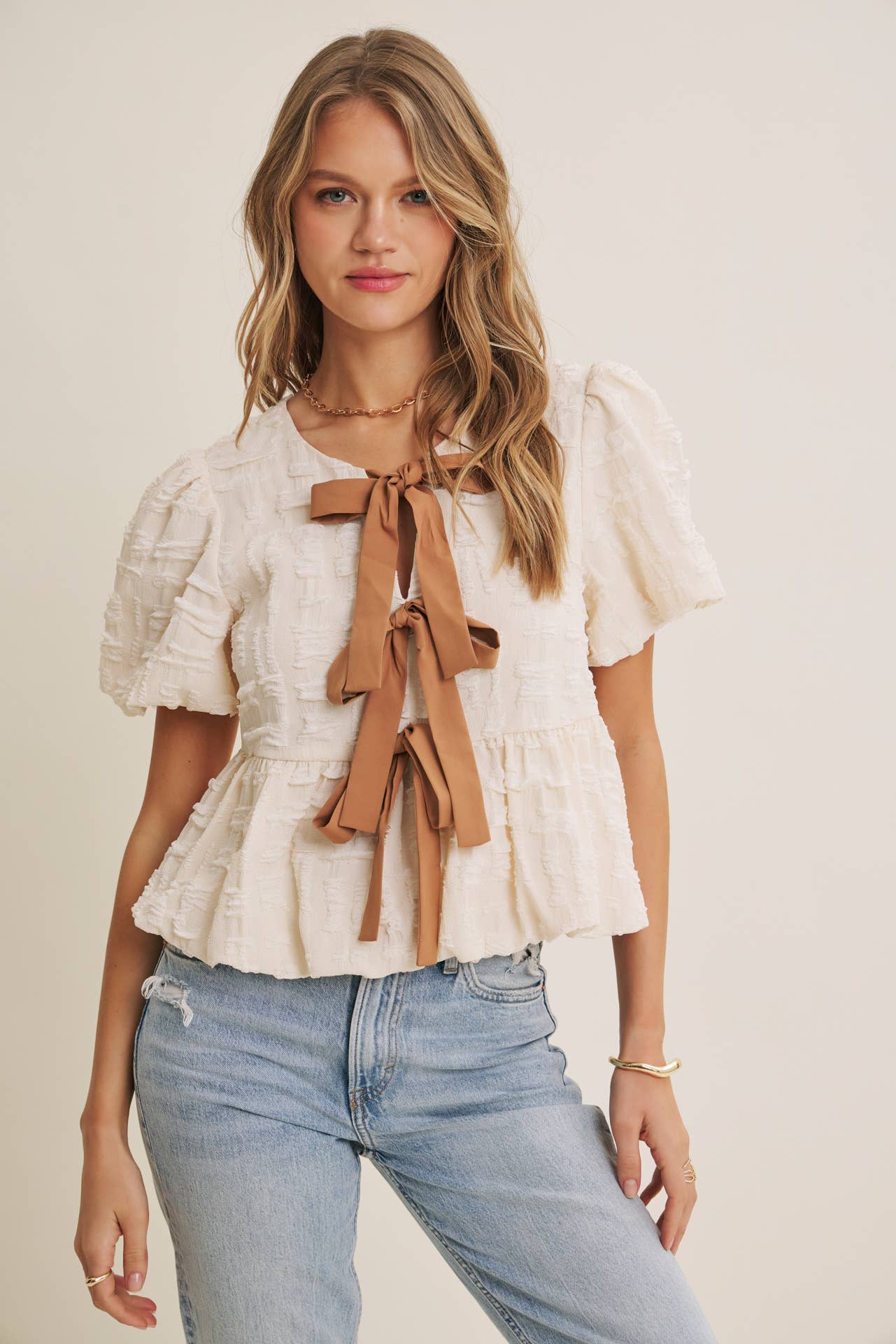 Woman wearing a white blouse with ruffles and a brown bow, paired with light blue jeans on a plain background.