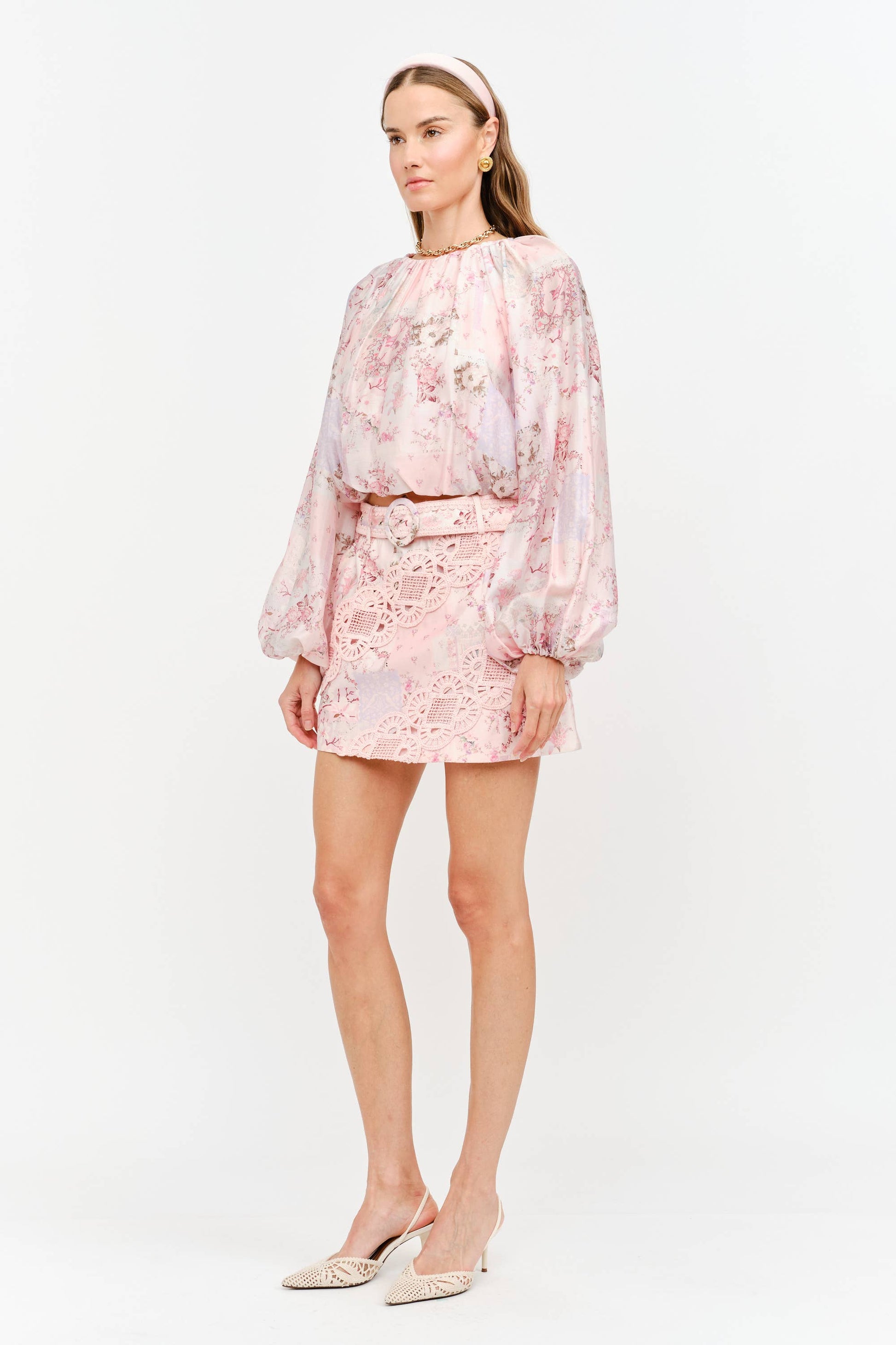 A model wearing a light pink floral lace two-piece set consisting of a long-sleeve top and a matching mini skirt, paired with beige pointed-toe heels against a plain white studio background.