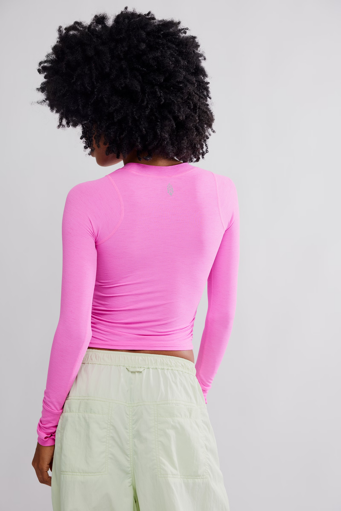 A woman with a large black afro is shown from the back wearing a bright pink long-sleeve crop top and light green pants against a plain light gray background.