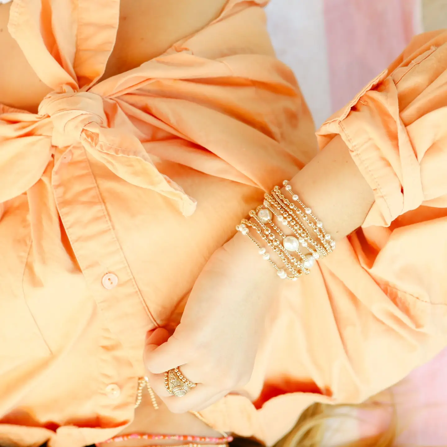 A close-up of a person wearing multiple stacked gold-toned bracelets with white pearl-like beads on one wrist, paired with a peach-colored blouse.