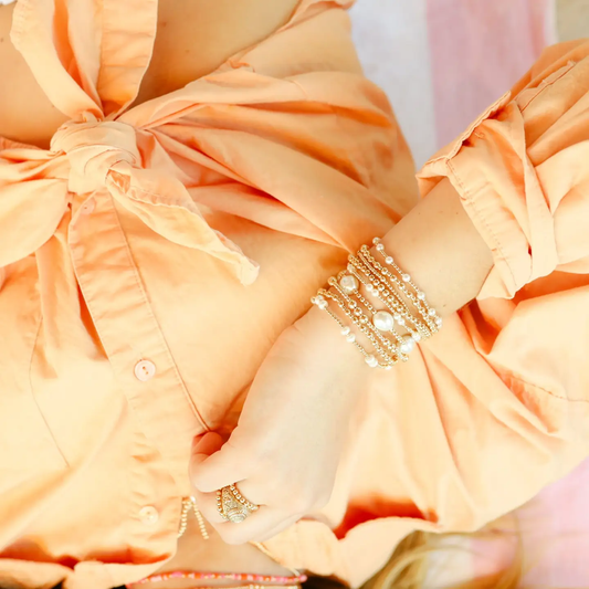 A close-up of a person wearing multiple stacked gold-toned bracelets with white pearl-like beads on one wrist, paired with a peach-colored blouse.