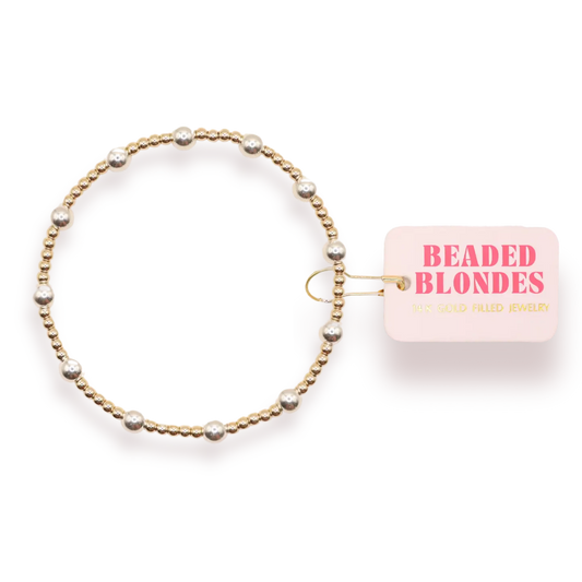 A gold beaded stretch bracelet with silver-toned pearl-like beads, shown next to a pink tag that reads 'BEADED BLONDES'.