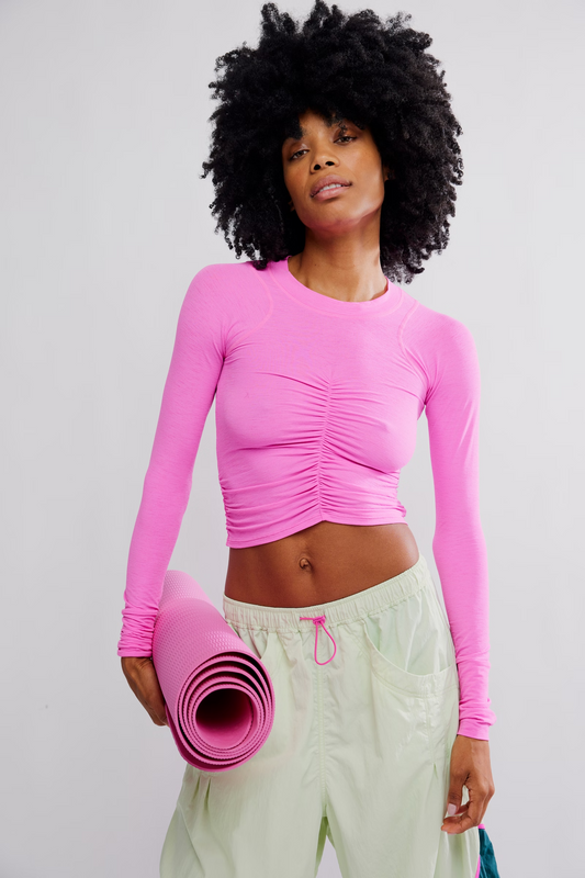 A woman with a large black afro hairstyle wearing a bright pink long-sleeve ruched crop top and light sage green yoga pants, holding a matching pink rolled-up yoga mat against a light gray studio background.