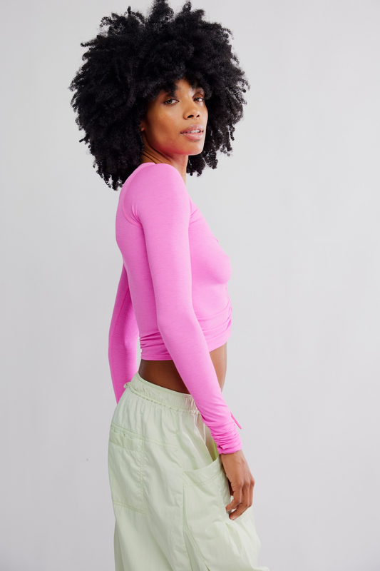 A woman with a large black afro is shown in profile wearing a bright pink long-sleeve crop top and light green pants against a plain light gray studio background.