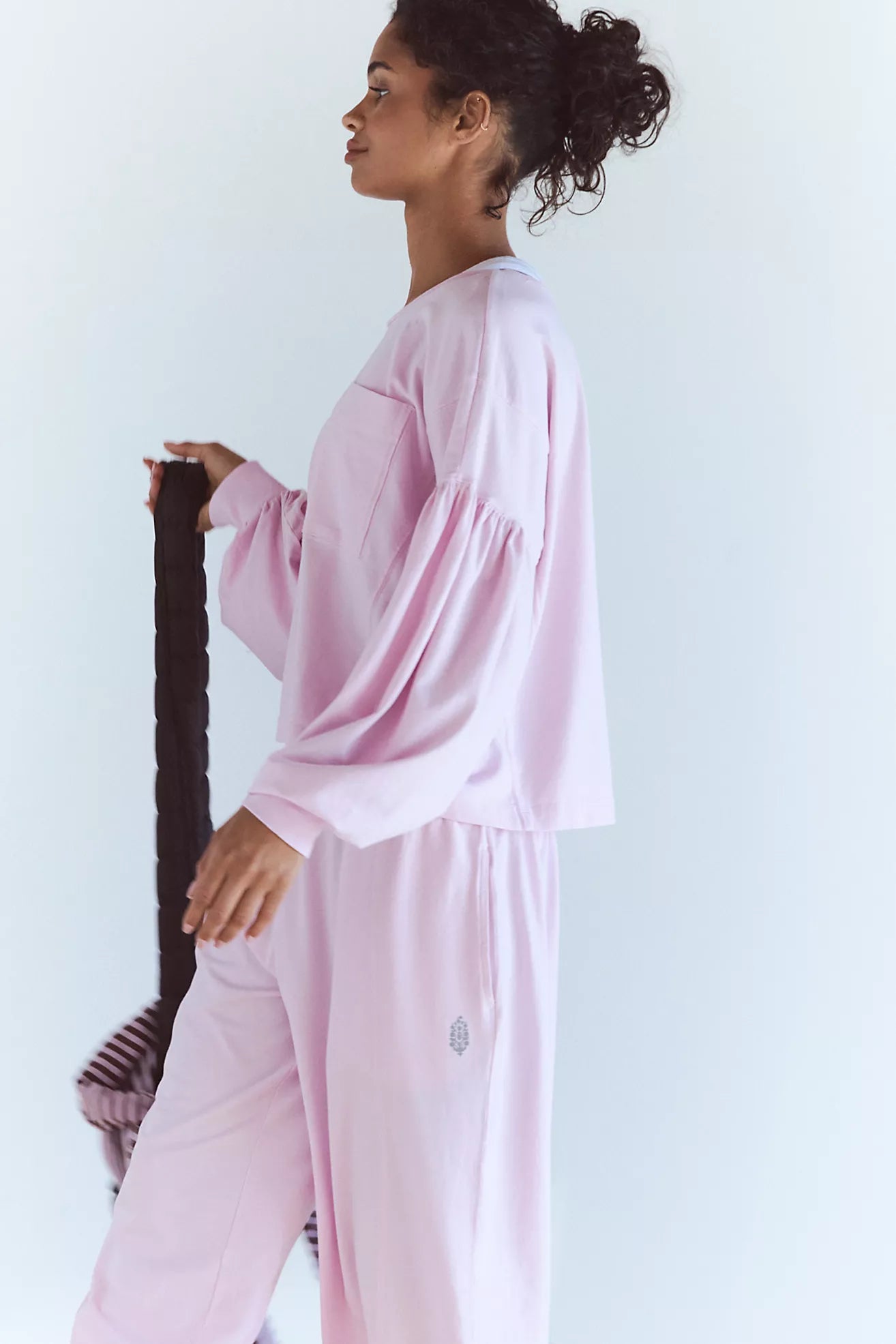 A woman wearing a light pink lounge set consisting of a long-sleeve top with voluminous balloon sleeves and matching wide-leg pants, holding a dark brown braided hair extension against a plain white background.