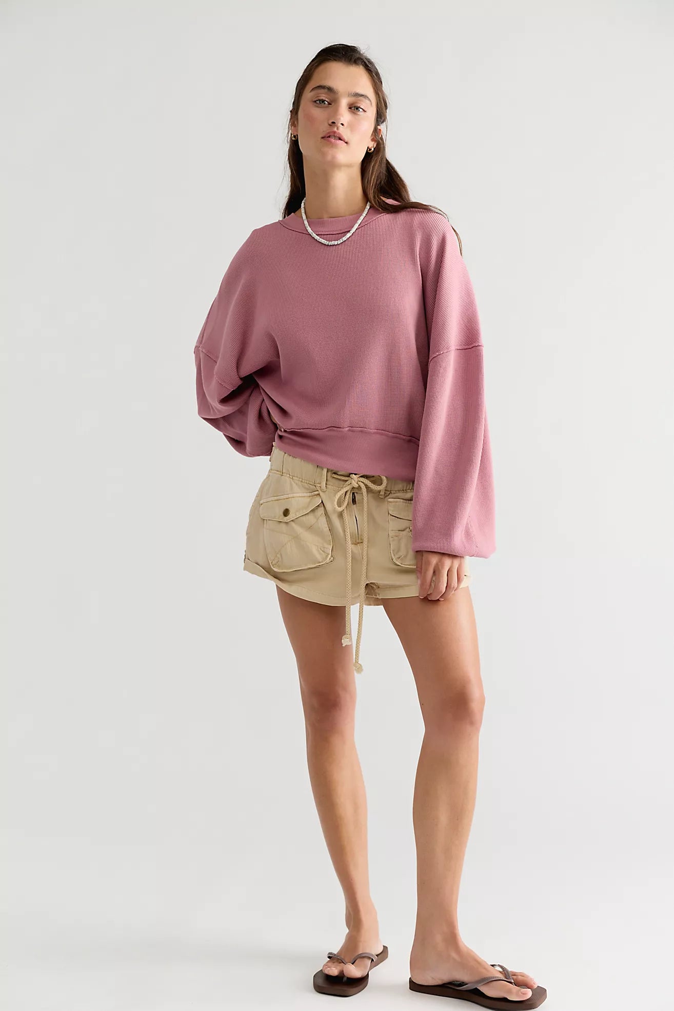 A woman modeling a dusty rose oversized long-sleeve sweatshirt and beige cargo shorts against a plain light gray studio background.