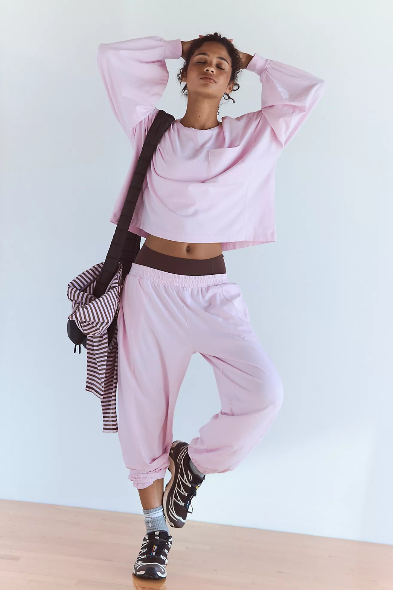 A woman modeling a light pink lounge set consisting of a cropped long-sleeve top and matching jogger pants, styled with a striped shirt draped over her shoulder and black sneakers.