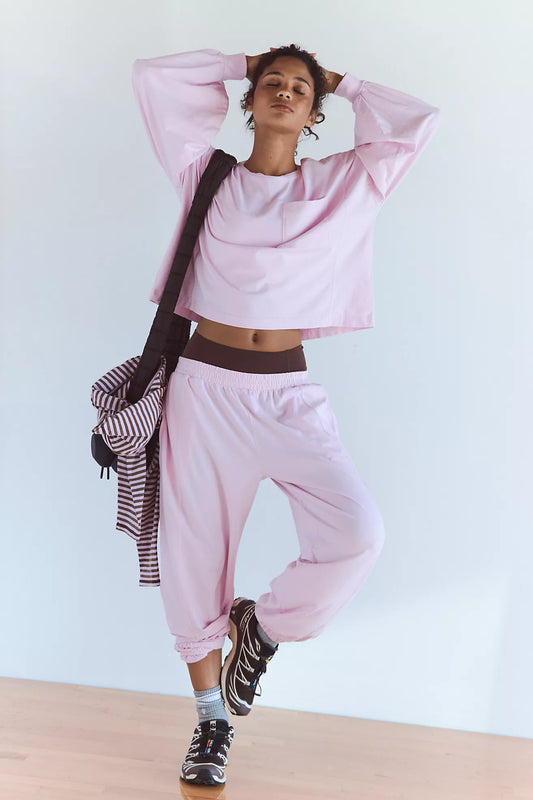 A woman modeling a light pink lounge set consisting of a cropped long-sleeve top and matching jogger pants, styled with a striped shirt draped over her shoulder and black sneakers.