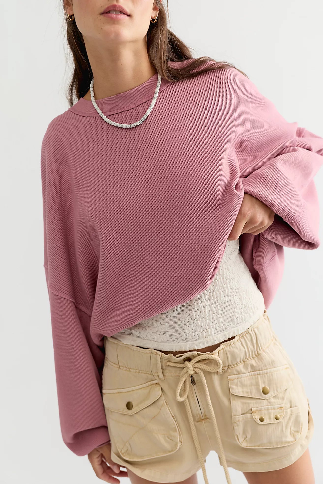 A woman wearing a dusty pink ribbed knit cropped sweatshirt, styled with a white textured top and beige drawstring shorts against a plain light background.