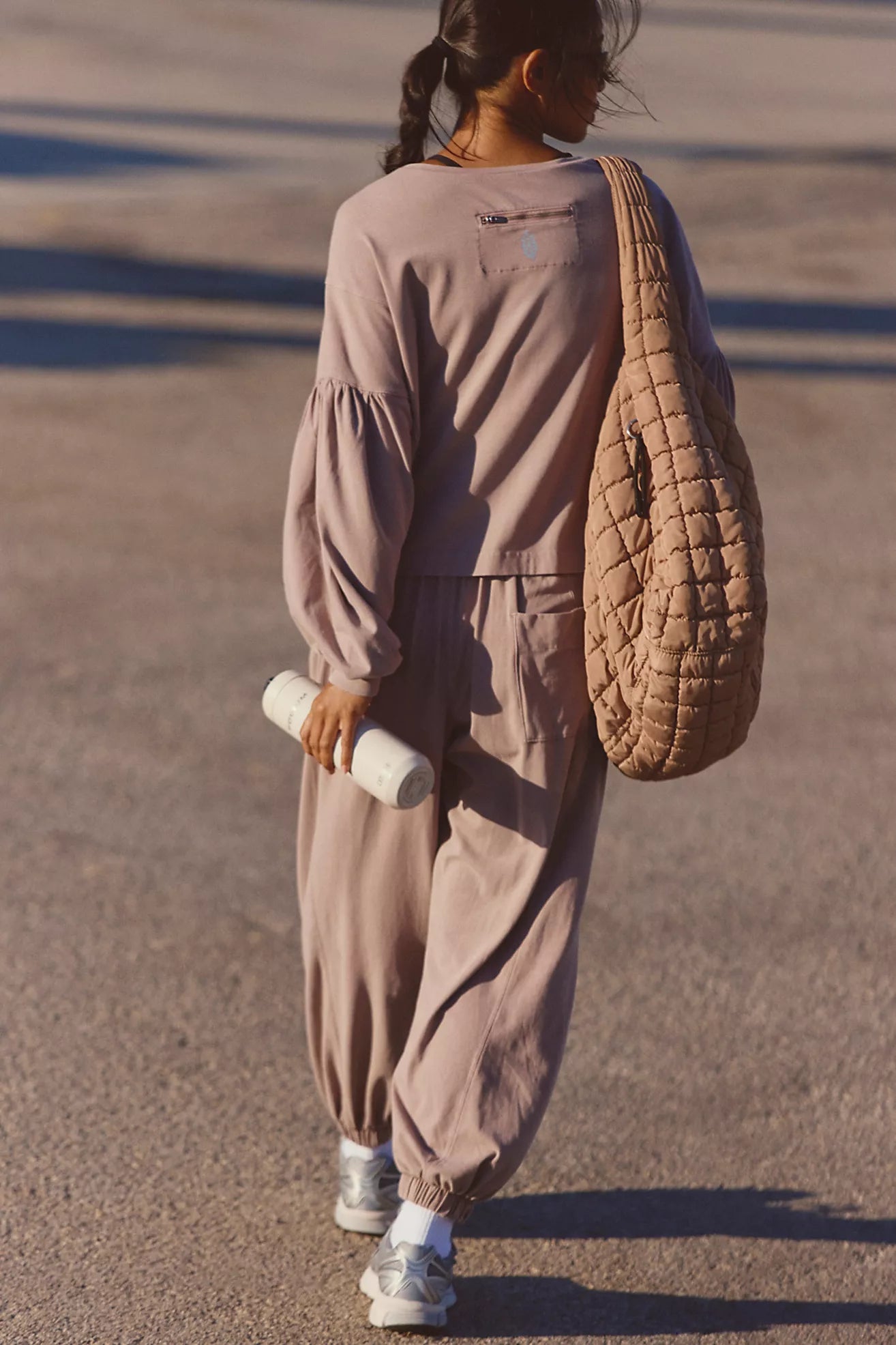 A person seen from behind wearing a matching dusty mauve two-piece lounge set with voluminous long sleeves and baggy jogger pants, carrying a large quilted tan shoulder bag and holding a white cylindrical item.