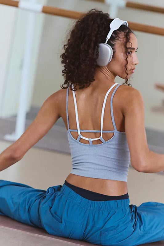 A woman in a yoga studio wearing a light blue-gray sports bra with white straps and matching blue pants, sitting in a split position and wearing white over-ear headphones.
